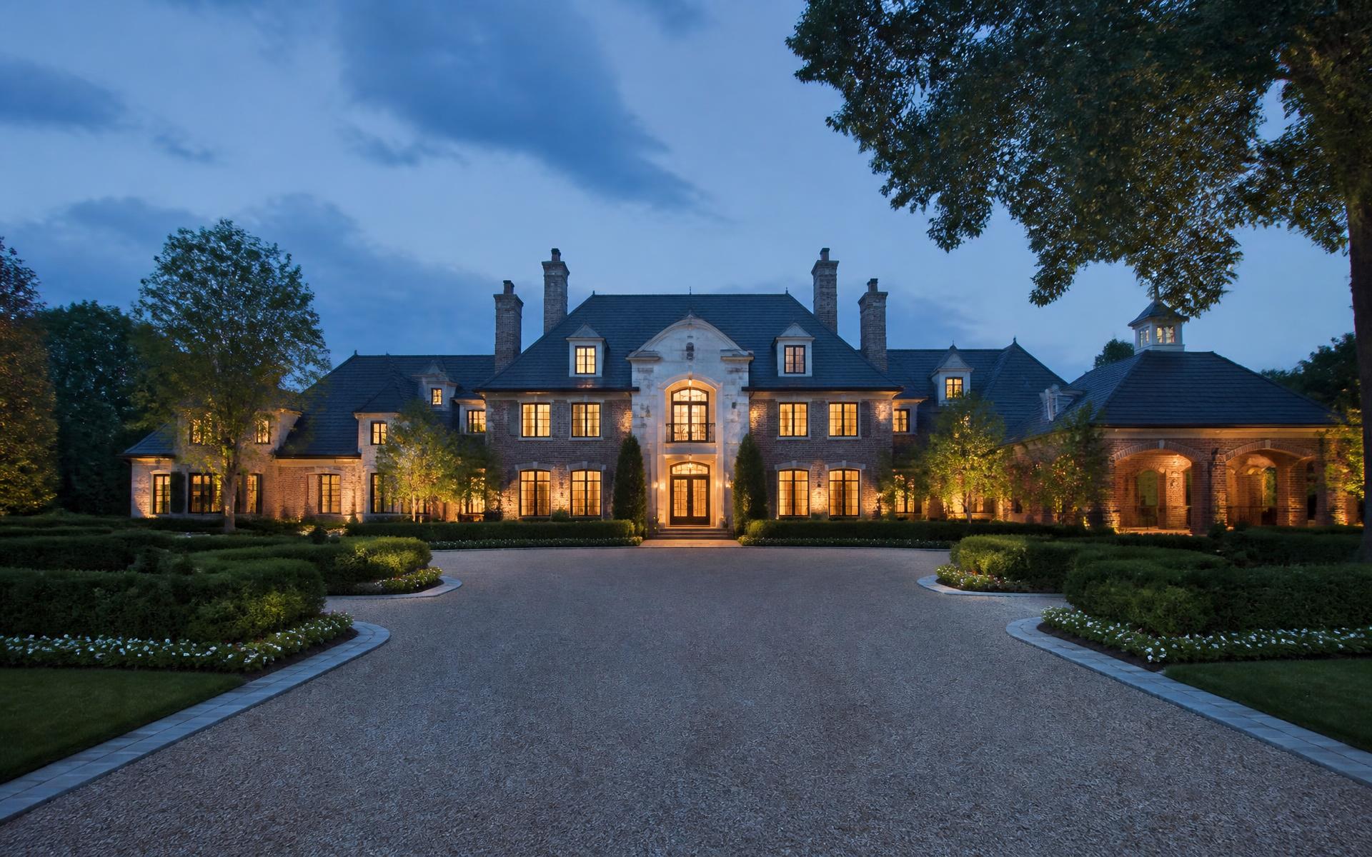 Illuminated brick and limestone estate house with formal gardens and a gravel motor court at dusk